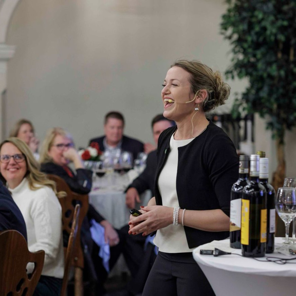 Claudia laughing as she hosts a wine tasting for a seated group, with bottles and a laptop beside her