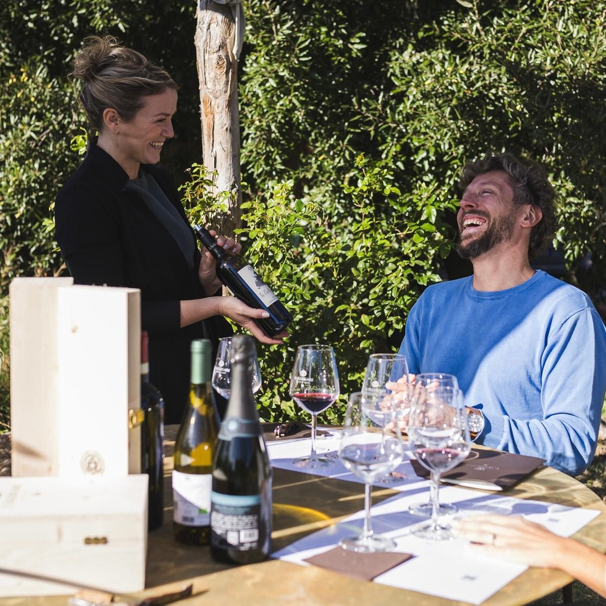 Claudia pouring red wine for a laughing guest at a garden table set for a tasting