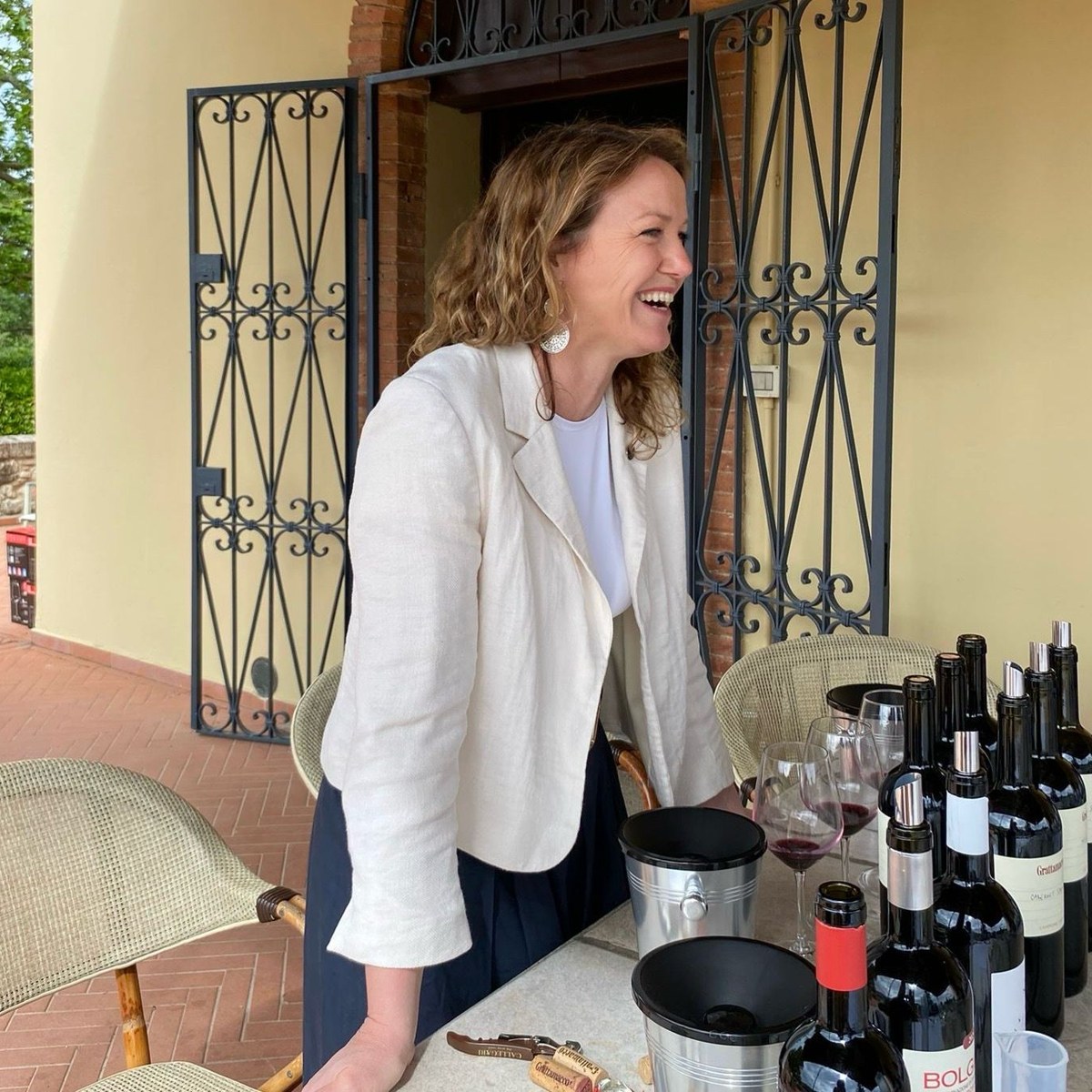Claudia laughing at a terrace table set with glasses and bottles of red wine for a tasting