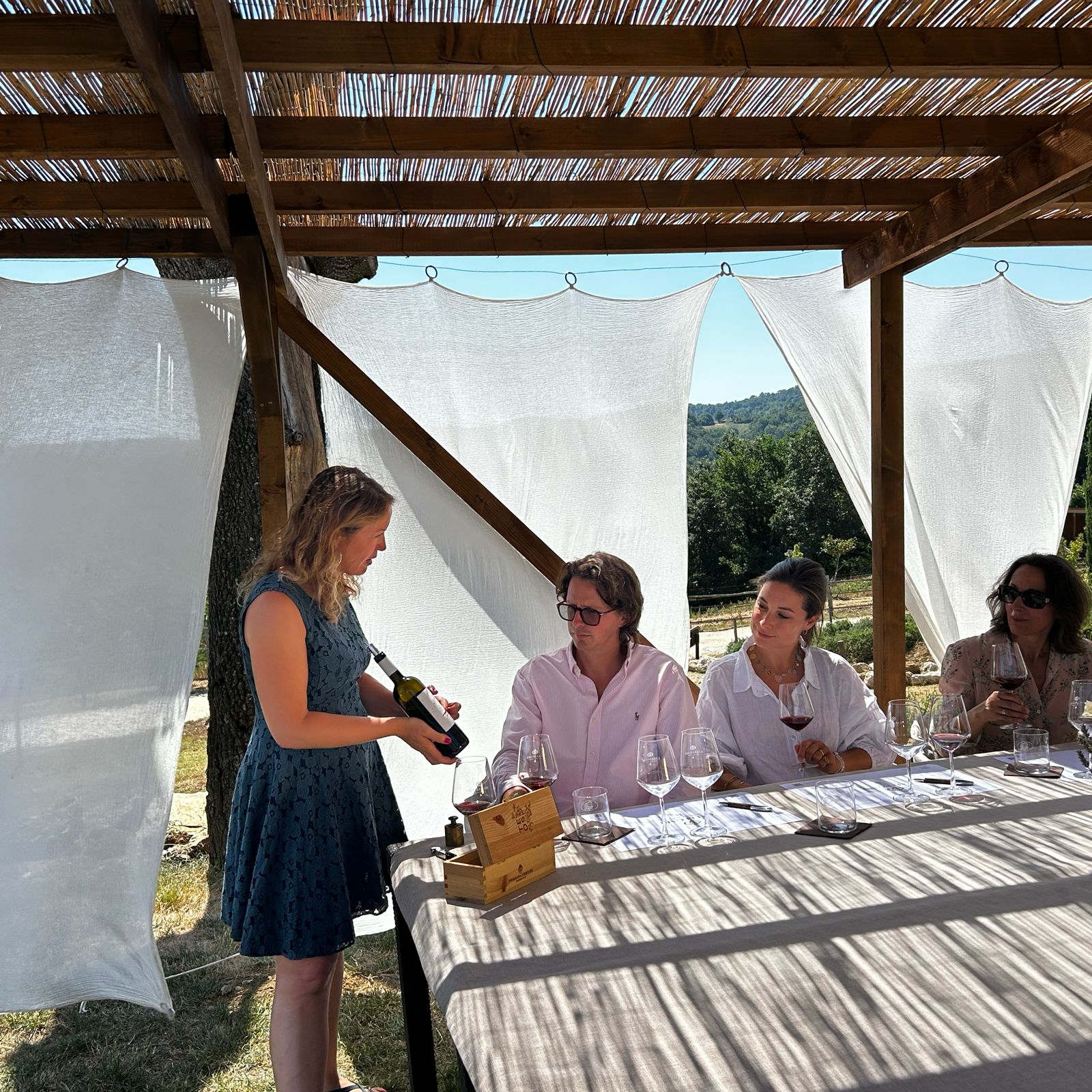 Claudia pouring red wine for guests at a private tasting under a pergola with a view of the Tuscan hills